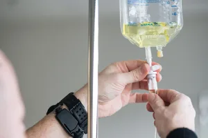 Evde damar yoluyla uygulama yapılırken sağlık personelinin serumu kontrol ettiği, hastanın kolunda serum setinin görüldüğü bir sahne.