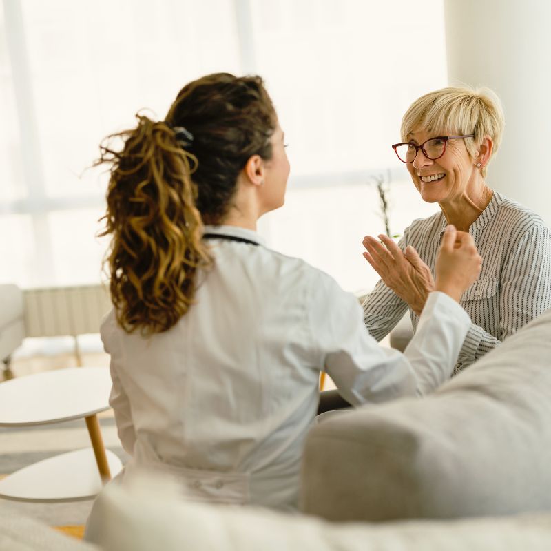 Ev ortamında doktorun hastayı dinlediği, hastanın rahat bir koltukta oturduğu bir muayene sahnesi.