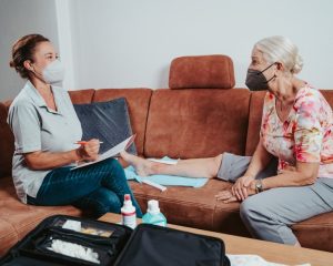 Evde muayenede doktorun hastayı dinleyip tansiyon ölçmesi, masada stetoskop ve not defterinin bulunması.