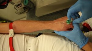 Evde sağlık personelinin hastadan kan aldığı ve serum hazırladığı, masada tıbbi malzemelerin bulunduğu bir sahne.