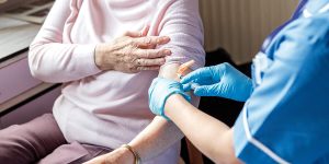 Evde sağlık hizmeti alan bir kişinin rahat bir ortamda dinlendiği, sağlık personelinin ölçüm yapıp not aldığı bir sahne.