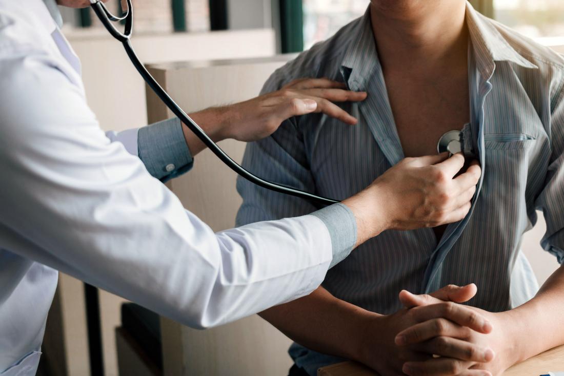 Evinin salonundaki koltuğunda oturan hastayı stetoskopla muayene eden, tıbbi ekipmanları yanında olan ve şikayetleri dikkatle dinleyen uzman doktor.